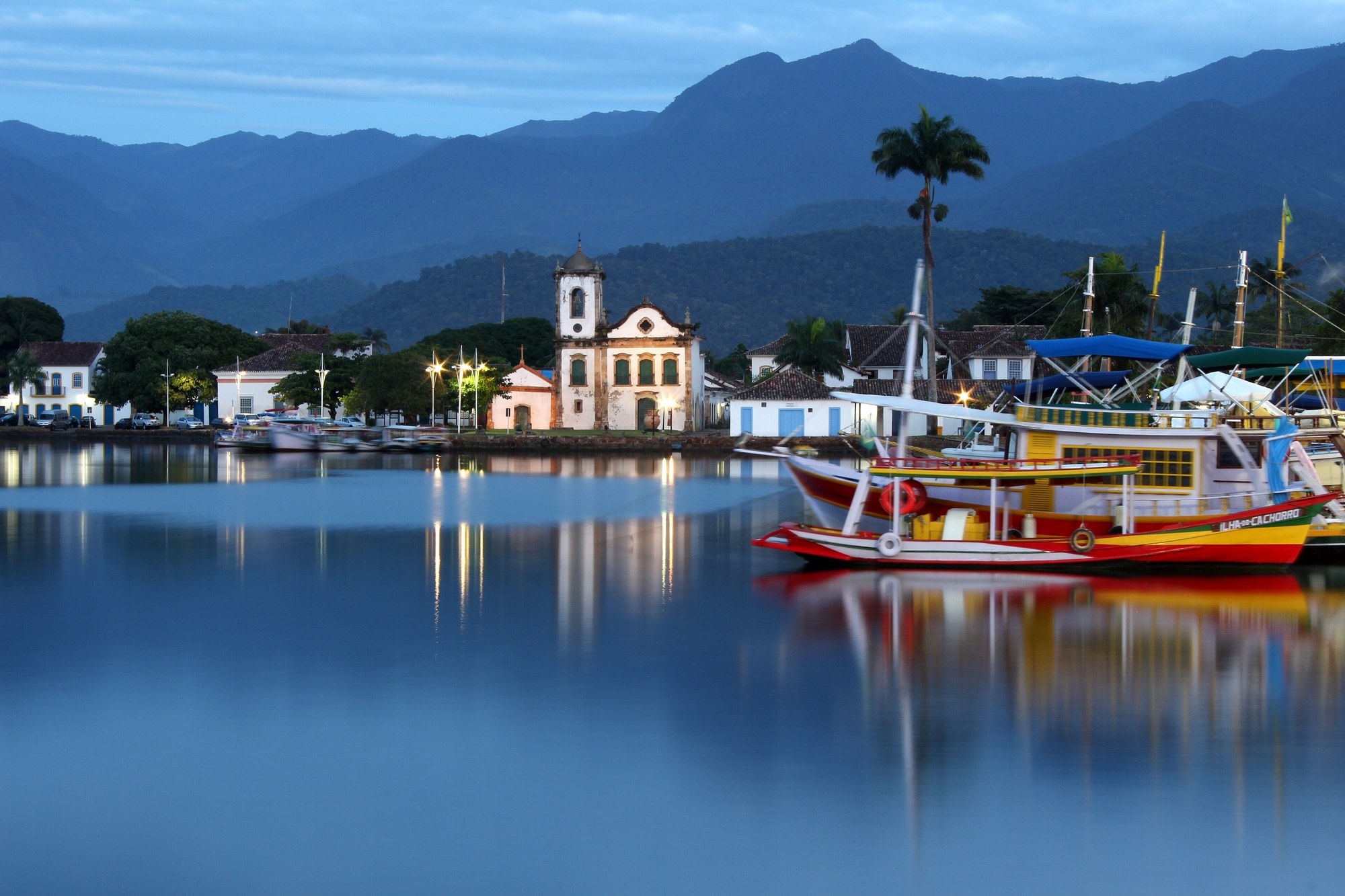 Igreja de Santa Rita à beira da baía de Paraty ao anoitecer, com barco iluminado.